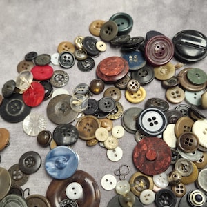 May include: A selection of assorted buttons in various colours, sizes, and materials. The buttons include black, brown, red, white, and blue, some with multiple holes and decorative patterns. The buttons are arranged on a grey surface.