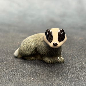 May include: A small ceramic badger figurine featuring a glossy finish. The animal has a cream-coloured face with distinct black markings around the eyes and ears, and a textured, grey-toned body with a white-tipped tail, captured in a resting pose.