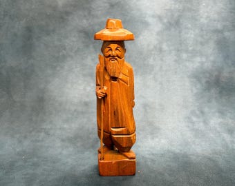May include: A hand-carved wooden figurine of an elderly person with a long beard, wearing a wide-brimmed hat and traditional robes. The figure stands on a rectangular base and holds a tall, thin walking stick in the right hand, featuring a warm brown wood grain finish throughout the entire sculpture.
