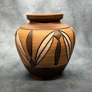 Peut inclure: Un vase de couleur terre cuite fait à la main avec une surface texturée et un design géométrique unique. Le vase présente un corps large et arrondi avec un col plus étroit et une base marron foncé. Le design comprend des formes de feuilles dans des tons de marron et de crème.