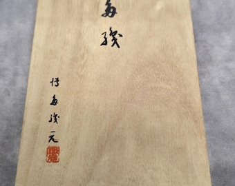 Puede incluir: Placa rectangular de madera clara con caligrafía japonesa en tinta negra. La veta de la madera es visible. Un sello rojo está en la parte inferior. El texto es una forma de arte tradicional japonés.