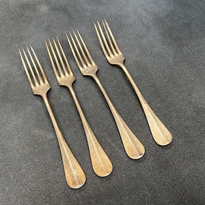 May include: A set of four matching metal forks with a tarnished brass-toned finish. Each fork features a slender handle that widens slightly at the base and four long, thin tines. The forks are arranged in a parallel diagonal row.