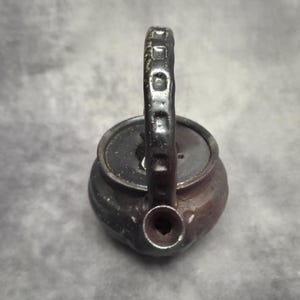 Puede incluir: Una pequeña tetera de estilo antiguo con un acabado oscuro y texturizado. El asa es de un color más oscuro y tiene un diseño rectangular. El pico es redondo y la tapa es plana. La tetera es probablemente de metal.