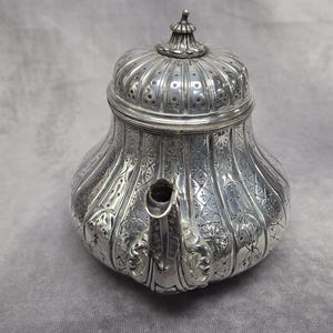 Op de afbeelding: Een sierlijke zilveren theepot met een gedetailleerd, in reliëf aangebracht bloemmotief. De theepot heeft een bolvormige body, een gebogen tuit en een gewelfde deksel met een decoratieve knop. Het oppervlak is zeer reflecterend en toont het ingewikkelde ontwerp.
