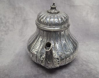 May include: Ornate silver teapot with a detailed, embossed floral pattern. The teapot has a bulbous body, a curved spout, and a domed lid with a decorative finial. The surface is highly reflective, showcasing the intricate design.