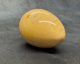 Puede incluir: Una piedra pulida y lisa con forma de huevo en un tono amarillo pálido cálido. La superficie presenta variaciones naturales sutiles, incluyendo tenues motas más oscuras y suaves vetas minerales horizontales en toda la forma.
