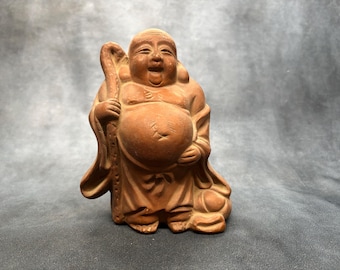 Puede incluir: Una figura de cerámica color terracota de un personaje sonriente y corpulento con la cabeza calva, vistiendo túnicas sueltas. El personaje sostiene un bastón alto y texturizado en la mano derecha y tiene un vientre grande y expuesto.