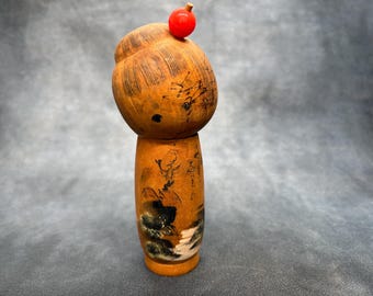 May include: A wooden Japanese Kokeshi doll with a rounded head and cylindrical body. The doll is painted with a light brown finish, with black ink details and a red ball on top of the head. The doll has a traditional design.