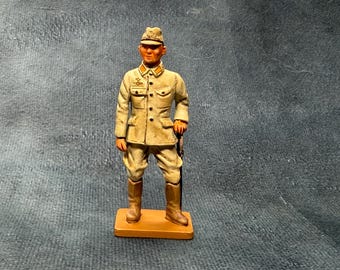 May include: A hand-painted miniature figurine of a soldier wearing a tan uniform, a matching peaked cap, and tall brown boots. The soldier stands on a rectangular tan base, with a small emblem on the chest and yellow collar tabs.