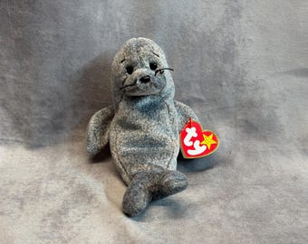 May include: A gray seal plush toy with a mottled texture, black plastic eyes, and stitched whiskers. The stuffed animal features a heart-shaped red and yellow Ty tag attached to its side, displaying the white letters ty and a yellow star.