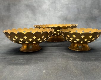 May include: Three gold-colored metal bowls with a pedestal base and a scalloped rim. Each bowl features a repeating pattern of small, cross-shaped cutouts along the sides, creating a perforated design that allows light to pass through the metal.