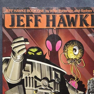May include: A Jeff Hawke Book One graphic novel cover featuring the text JEFF HAWKE BOOK ONE by Willie Patterson and Sydney Jordan. The artwork displays a large metallic robot figure, a floating yellow tentacled creature, and a man in a uniform.