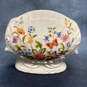 May include: White ceramic shell-shaped decorative bowl with a floral and butterfly design. The bowl has a scalloped edge and a pedestal base. The floral design includes red, blue, yellow, and pink flowers with green leaves.