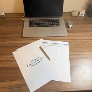 May include: A laptop, watch, and documents are on a wooden desk. The documents include a "Weekly Performance Planner" and "Annual Performance Vision". A silver pen rests on the documents. The laptop is open, displaying a black screen.