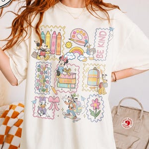 May include: An ivory t-shirt featuring a graphic print of Disney characters including Mickey, Minnie, Donald, Daisy, Goofy, and Pluto. The design includes school-themed elements like crayons, books, an alphabet chart, and the text BE KIND.