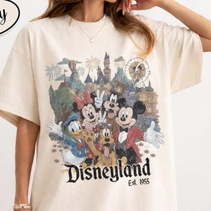Może przedstawiać: Kremowa koszulka z grafiką Disneyland z Myszką Miki, Minnie, Kaczorem Donaldem, Goofym i Pluto. Grafika zawiera zamek, fajerwerki i diabelski młyn. Tekst "Disneyland Est. 1955" jest wydrukowany poniżej.