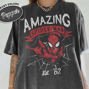 May include: Dark grey t-shirt featuring a distressed graphic of Spider-Man with the text "The Amazing Spider-Man" and "Est. '62" in red and white. The shirt has a vintage, worn look and a relaxed fit. The brand name "Pepper" is in a circle on the top left.