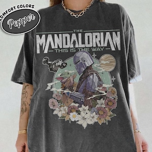 May include: Dark gray t-shirt featuring a graphic of The Mandalorian and Grogu surrounded by flowers. The text "THE MANDALORIAN - THIS IS THE WAY" is printed above the image. The shirt has a Comfort Colors Pepper label.