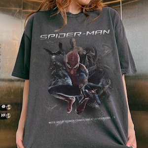 May include: Dark gray Spider-Man t-shirt featuring a graphic of the superhero in a dynamic pose, surrounded by other characters. The text "SPIDER-MAN" is displayed above the image, with the phrase "WITH GREAT POWER COMES GREAT RESPONSIBILITY" below.