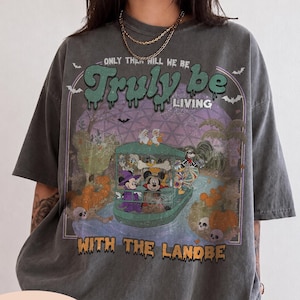 May include: A charcoal gray t-shirt featuring a graphic print of cartoon characters in a boat on a river. The text reads "ONLY THEN WILL WE BE TRULY BE LIVING WITH THE LANDBE". The design includes pumpkins, bats, and a purple sky background.