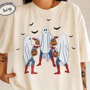 May include: Ivory-colored t-shirt featuring a Halloween-themed design. Three figures dressed as ghosts are holding jack-o'-lantern buckets. The figures have Spider-Man-inspired legs and are surrounded by black bat silhouettes. The shirt has the words "Comfort Colors" and "Ivory" in a circle.