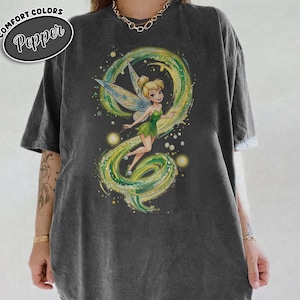 May include: Dark grey t-shirt with a graphic of Tinkerbell, featuring green and yellow details. The fairy is set against a swirling design. The shirt includes the words "Comfort Colors" and "Pepper" in a circular design.