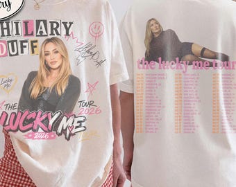 Könnte beinhalten: Ein elfenbeinfarbenes Kurzarm-T-Shirt mit einer Grafik von Hilary Duff auf der Vorderseite, inklusive des Textes "HILARY DUFF", "THE LUCKY ME TOUR 2026" und einer Unterschrift. Die Rückseite zeigt ein Foto von Hilary Duff und den Text "the lucky me tour" mit einer Liste von Daten.