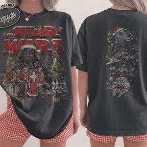 Könnte beinhalten: Ein anthrazitfarbenes T-Shirt mit einem Star Wars-Weihnachtsmotiv. Die Vorderseite zeigt Darth Vader, Chewbacca und weitere Figuren mit Weihnachtsmützen vor einem Tannenbaum. Die Rückseite zeigt eine vertikale Anordnung des Todessterns, eines X-Wings und des Millennium Falken mit weihnachtlichen Dekorationen.