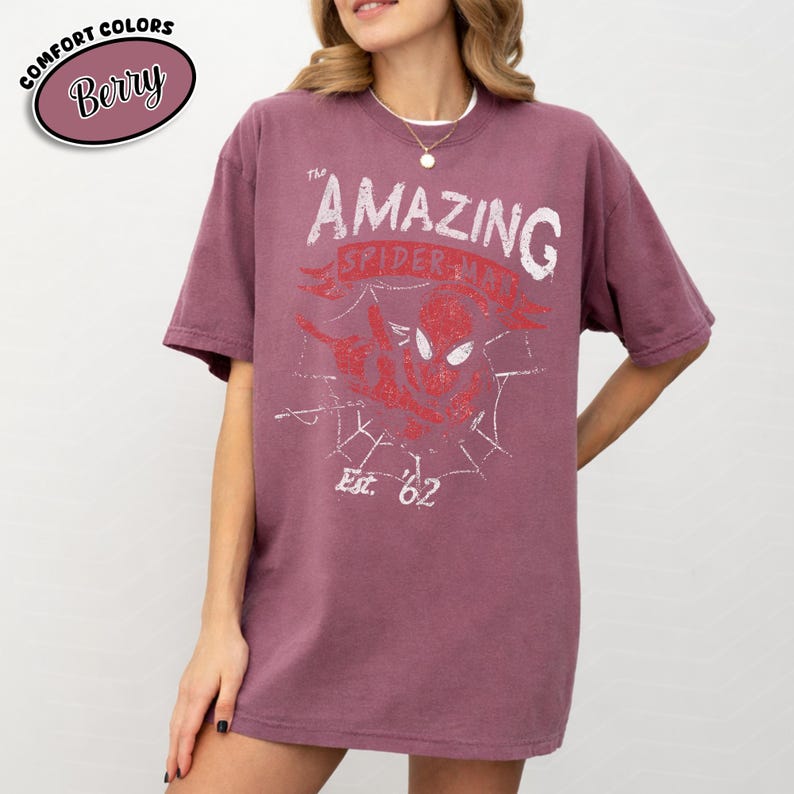 May include: Berry-colored t-shirt with a distressed graphic of Spider-Man and the text "The Amazing Spider-Man Est. '62". The shirt has short sleeves and a relaxed fit. The words "Comfort Colors" and "Berry" are in a circle in the upper left corner.