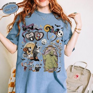 May include: A blue short-sleeve t-shirt featuring a graphic print of characters from The Nightmare Before Christmas, including Jack Skellington, Sally, and Oogie Boogie. The design includes a purple bow, spiderwebs, and the text Nightmare.