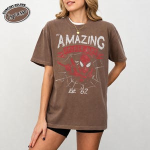 May include: Espresso-colored t-shirt featuring a distressed graphic of Spider-Man with the text "THE AMAZING SPIDER-MAN" and "Est. '62". The shirt has a vintage look and feel.