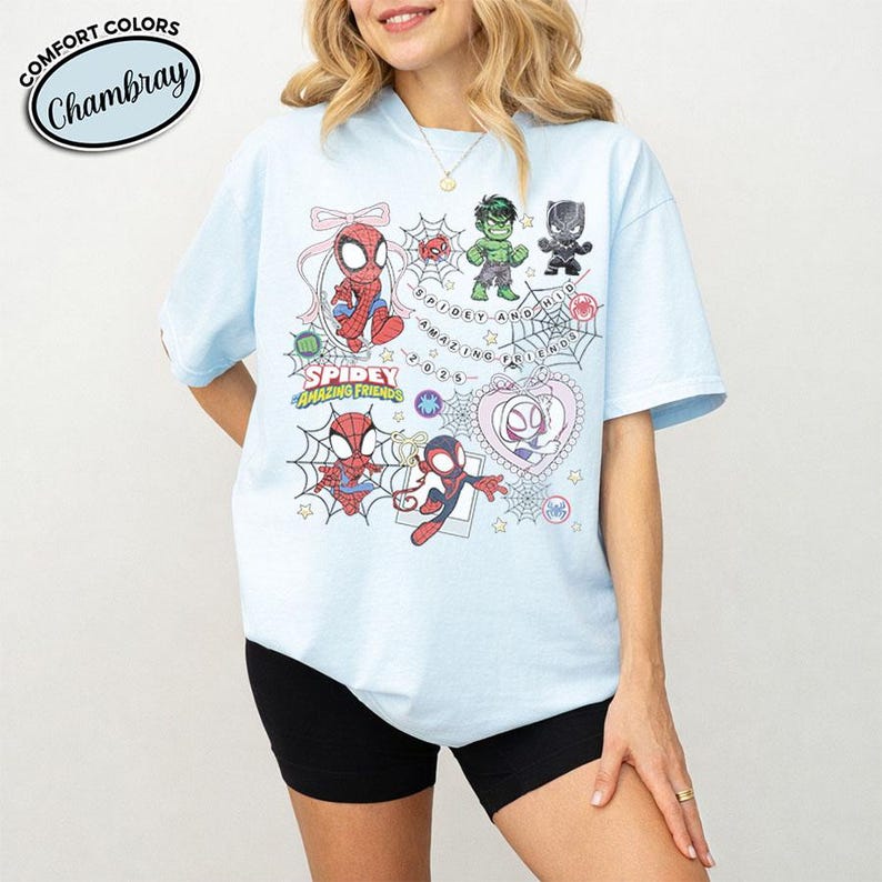 以下が含まれることがあります： スパイダーマンなどのキャラクターの漫画イラストが描かれた水色のTシャツ。「Spidey and Amazing Friends」と「Comfort Colors Chambray」の文字が円形にデザインされています。