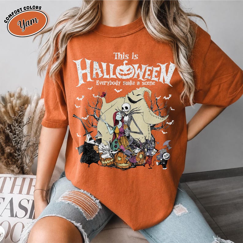 Puede incluir: Camiseta naranja con un dise&ntilde;o de "Pesadilla antes de Navidad". El dise&ntilde;o presenta a Jack Skellington, Sally y otros personajes, con el texto "This is Halloween". La camiseta tiene mangas cortas y cuello redondo.