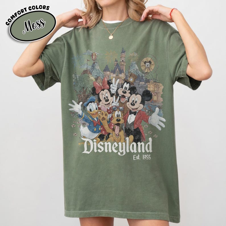 Może przedstawiać: Zielona koszulka w kolorze mchu z grafiką Disneyland w stylu vintage. Projekt zawiera Myszkę Miki, Minnie, Kaczora Donalda, Psa Pluto i inne postacie. Tekst "Disneyland Est. 1955" jest wydrukowany poniżej. Koszulka ma kr&oacute;tkie rękawy i okrągły dekolt.
