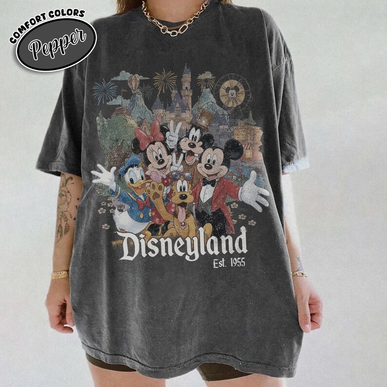 Może przedstawiać: Ciemnoszara koszulka z grafiką Disneyland z Myszką Miki, Minnie, Kaczorem Donaldem, Goofym i innymi postaciami. Koszulka ma napis "Disneyland Est. 1955" i logo Comfort Colors Pepper.