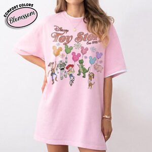 K&ouml;nnte beinhalten: Hellrosa T-Shirt mit dem Text "Disney Toy Story Est. 1995" und Zeichentrickfiguren, die Mickey Mouse-f&ouml;rmige Ballons halten. Das Shirt hat wei&szlig;e Abschl&uuml;sse an den &Auml;rmeln und den Text "Comfort Colors Blosssom" in einem rosa Oval.