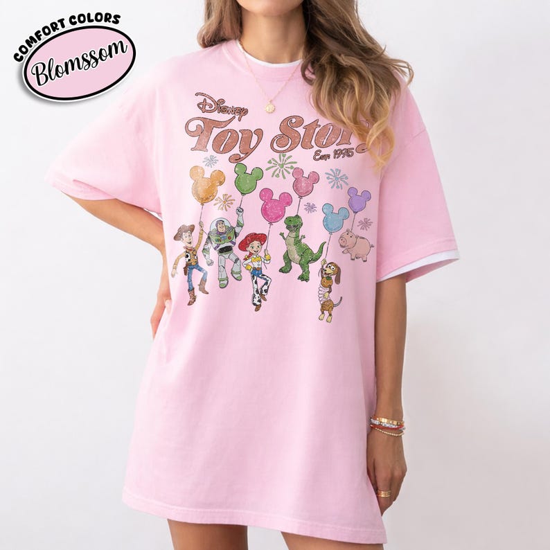 K&ouml;nnte beinhalten: Hellrosa T-Shirt mit dem Text "Disney Toy Story Est. 1995" und Zeichentrickfiguren, die Mickey Mouse-f&ouml;rmige Ballons halten. Das Shirt hat wei&szlig;e Abschl&uuml;sse an den &Auml;rmeln und den Text "Comfort Colors Blosssom" in einem rosa Oval.