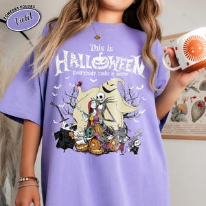 Puede incluir: Una camiseta violeta con un dise&ntilde;o gr&aacute;fico de "Pesadilla antes de Navidad". El dise&ntilde;o presenta a Jack Skellington, Sally y otros personajes, con el texto "This is Halloween" y "Everybody make a scene."