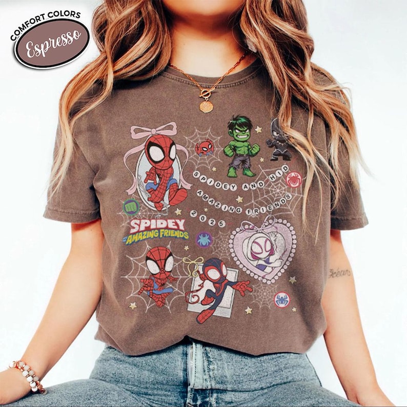 以下が含まれることがあります： 「スパイディとすごい仲間たち」のキャラクター、スパイダーマン、ハルクなどの漫画イラストが描かれたエスプレッソカラーのTシャツ。「Spidey Amazing Friends」の文字とその他のグラフィックがプリントされています。