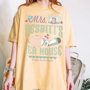 K&ouml;nnte beinhalten: Senfgelbes T-Shirt mit einem skurrilen Design. Die Grafik zeigt "Mrs. Nesbitt's Tea House" mit einer Teekanne, einer Figur und dem Satz "To Infini-tea and Beyond". Das Design ist in Gr&uuml;n-, Rosa- und Lilat&ouml;nen gehalten.