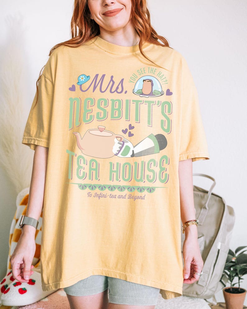 K&ouml;nnte beinhalten: Senfgelbes T-Shirt mit einem skurrilen Design. Die Grafik zeigt "Mrs. Nesbitt's Tea House" mit einer Teekanne, einer Figur und dem Satz "To Infini-tea and Beyond". Das Design ist in Gr&uuml;n-, Rosa- und Lilat&ouml;nen gehalten.