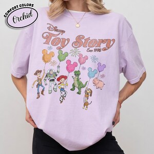 K&ouml;nnte beinhalten: Helllila Comfort Colors T-Shirt mit Toy Story-Grafik. Das Design zeigt Charaktere, die Mickey Mouse-Ballons halten, mit dem Text "Disney Toy Story Est. 1995". Das T-Shirt hat eine lockere Passform.