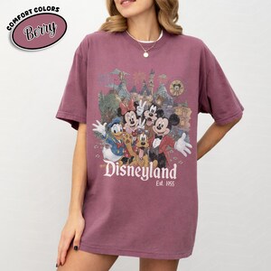 Może przedstawiać: Koszulka w kolorze jagodowym z grafiką Disneyland z Myszka Miki, Myszka Minnie, Kaczorem Donaldem, Goofym i Plutonem. Grafika zawiera zamek i napis "Disneyland Est. 1955". Koszulka ma kr&oacute;tkie rękawy i okrągły dekolt.