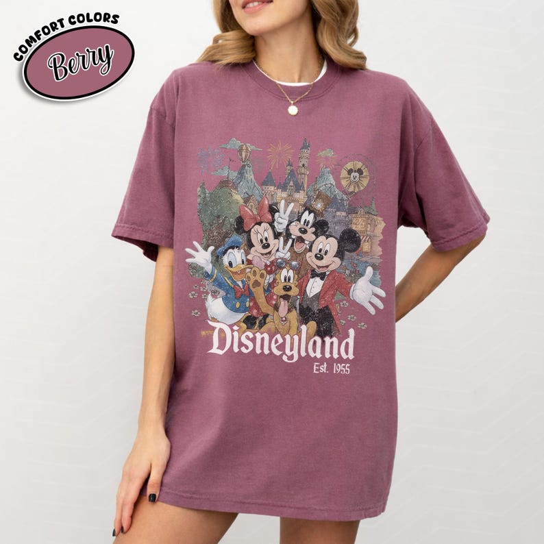 Może przedstawiać: Koszulka w kolorze jagodowym z grafiką Disneyland z Myszka Miki, Myszka Minnie, Kaczorem Donaldem, Goofym i Plutonem. Grafika zawiera zamek i napis "Disneyland Est. 1955". Koszulka ma kr&oacute;tkie rękawy i okrągły dekolt.