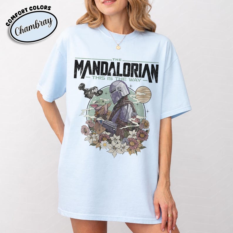 May include: Light blue t-shirt featuring a graphic of The Mandalorian and Grogu, surrounded by flowers, with the text "THE MANDALORIAN - THIS IS THE WAY." The shirt has a relaxed fit and the text "Comfort Colors Chambray" in a circle.