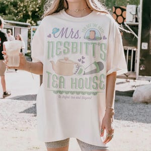 K&ouml;nnte beinhalten: Cremefarbenes T-Shirt mit dem Text "Mrs. Nesbitt's Tea House" in Gr&uuml;n und Lila. Das Design umfasst eine Teekanne, Herzen und den Satz "To Infini-tea and Beyond". Das Shirt hat kurze &Auml;rmel.