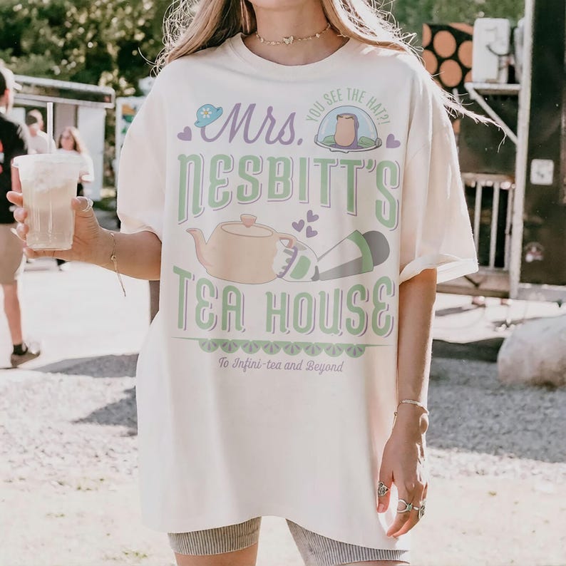 K&ouml;nnte beinhalten: Cremefarbenes T-Shirt mit dem Text "Mrs. Nesbitt's Tea House" in Gr&uuml;n und Lila. Das Design umfasst eine Teekanne, Herzen und den Satz "To Infini-tea and Beyond". Das Shirt hat kurze &Auml;rmel.
