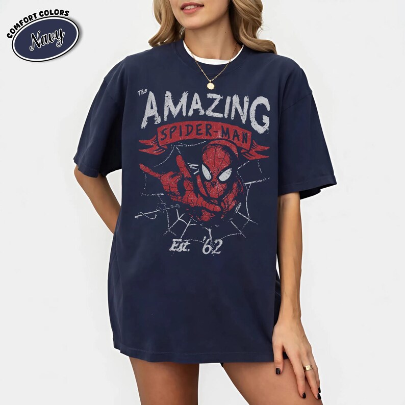 May include: Navy blue t-shirt featuring a distressed graphic of Spider-Man with the text "THE AMAZING SPIDER-MAN" and "Est. '62". The graphic is in red and white. The shirt has a relaxed fit and short sleeves.
