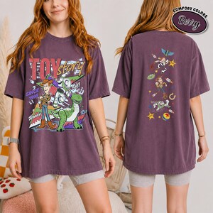 K&ouml;nnte beinhalten: Ein beerenfarbenes Kurzarm-T-Shirt mit Toy Story-Grafiken. Die Vorderseite zeigt Woody, Buzz Lightyear und Rex mit dem Text Toy Story Since 1995. Die R&uuml;ckseite zeigt eine vertikale Anordnung von Charakteren, darunter Woody und Buzz.