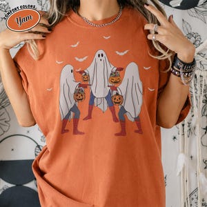 May include: Orange Comfort Colors t-shirt with a Halloween-themed design. The graphic depicts three ghosts dressed as superheroes, carrying pumpkin baskets, with bats overhead. The shirt has a relaxed fit.